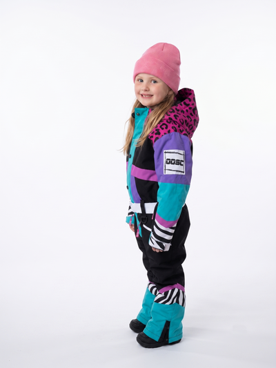 A young girl modeling a colorful winter ski suit with a pink beanie, standing against a plain white studio background. The ski suit features a color-blocked design of black, purple, teal, and pink with leopard and zebra print details.